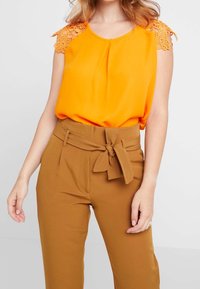 Blusa senza maniche di un luminoso arancione, con dettagli in pizzo sulle spalle, abbinata a pantaloni marroni a vita alta con una cintura legata.