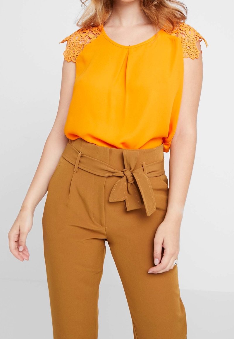 Blusa senza maniche di un luminoso arancione, con dettagli in pizzo sulle spalle, abbinata a pantaloni marroni a vita alta con una cintura legata.