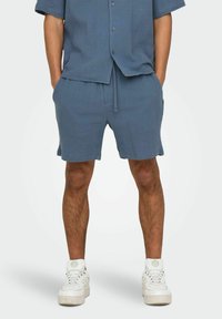 Lichtblauwe shorts met korte mouwen en bijpassende shorts, textuurstof, trekkoord in de taille, gecombineerd met witte sneakers. Eenvoudige knoopdetails.