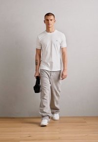 Maglietta bianca a maniche corte, pantaloni grigi comodi e scarpe da ginnastica bianche. Porta un oggetto nero, con una texture liscia e un design minimal.