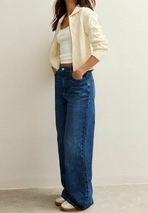 Femme portant un cardigan crème en maille torsadée sur un débardeur blanc, associé à un jean bleu taille haute à jambes larges et des chaussures blanches, les mains dans les poches.