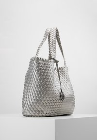 Borsa intrecciata argento con manici doppi, caratterizzata da una superficie testurizzata, forma arrotondata e un ciondolo logo circolare attaccato a un lato.