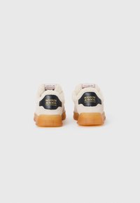 Sneakers beige con un morbido collo bianco, logo del brand nero e oro sul retro e suola in gomma trasparente arancione.