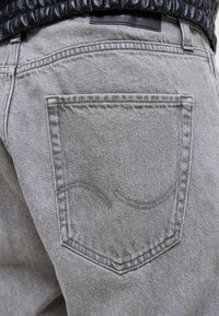 Primo piano di una persona che indossa jeans denim grigio chiaro con un disegno ricamato curvo sulla tasca posteriore e una fascia nera della giacca.