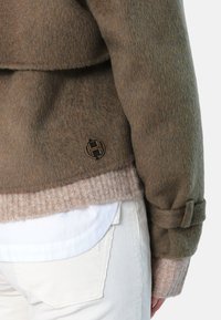 Strikket brun ulljakke med beige ribbede mansjetter, med en rund sort og gull logo-detalj på siden. Glatt, teksturert stoff.