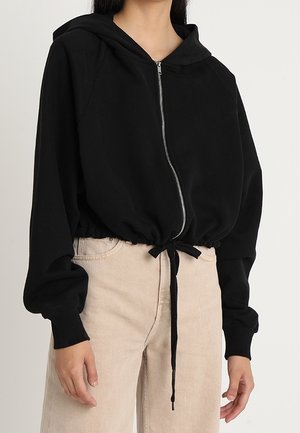 Kobieta ubrana w czarną bluzę z suwakiem i ściągaczem w talii, zestawioną z jasnobeżowymi, wysokim stanem spodniami na jednolitym tle.