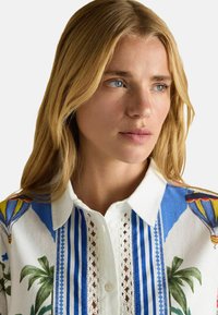 White collared shirt with blue and green floral pattern, featuring lace accents and vertical striped design. Buttons down the front.