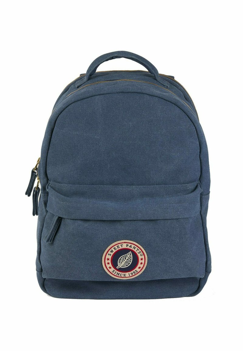 Sac à dos en toile marine avec un dessus arrondi, zips dorés, poche avant et un patch logo circulaire représentant une feuille, avec des accents rouges et blancs.