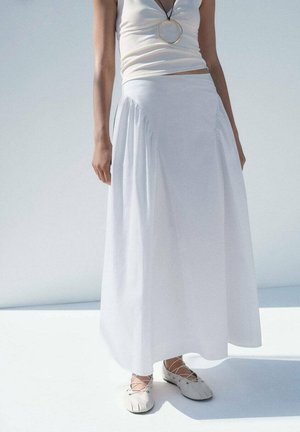 Femme portant un haut blanc sans manches, une longue jupe blanche, un collier avec pendentif rond et des ballerines blanches à lacets, debout sur une surface claire.