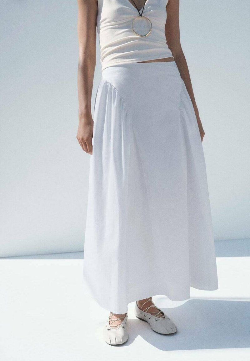 Femme portant un haut blanc sans manches, une longue jupe blanche, un collier avec pendentif rond et des ballerines blanches à lacets, debout sur une surface claire.