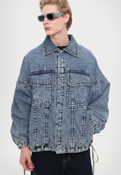 Jachetă din denim de culoare albastru deschis, cu o croială lejeră, având două buzunare frontale, cusături vizibile și manșete elastice cu detalii de cordon.