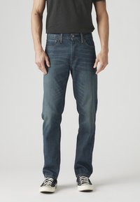 Homme portant un jean droit bleu foncé, un t-shirt noir et des baskets noires avec des lacets blancs, debout devant un fond uni.