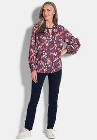 Bunt gemusterte Bluse mit Schlüsselloch-Ausschnitt, langen Ärmeln und einem Muster in den Farben Rosa, Lila und Grün, kombiniert mit dunklen Hosen.