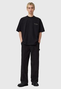 Zwarte oversized t-shirt met een klein logo op de borst, gecombineerd met donkere corduroy broek en zwarte schoenen, staand tegen een lichte achtergrond.