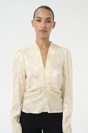 Blouse en satin crème avec des manches longues, décolleté en V profond et détails froncés à la taille. Présente un motif texturé subtil.