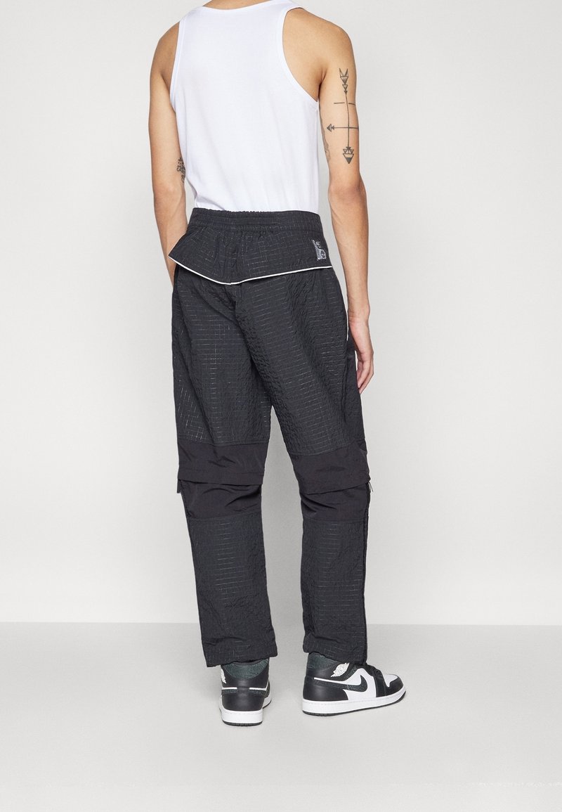 Pantalon noir avec un motif texturé et un tissu lisse contrastant sur le bas des jambes. Dispose d'une taille élastique et de fermetures éclair latérales.