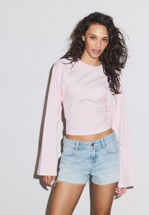 Femme aux longs cheveux bouclés portant un haut à manches longues rose clair et un short en jean bleu clair, debout devant un fond blanc uni.