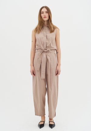 Beige ärmelloser Jumpsuit mit Kragen, tailliertem Schnitt, weitem Bein-Design und seitlichen Schlitzen. Kombiniert mit schwarzen spitzen Schuhen.