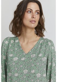Abito floreale verde con scollatura a V, tessuto texture e dettagliati motivi bianchi di fiori e foglie. Maniche lunghe con spalle arricciate.