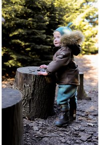 Child's brown hooded jacket with fur trim, teal pants, and black rubber boots. Standing beside a stump, textured ground with wood chips.