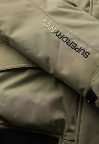 Oliwkowa kurtka o gładkiej fakturze, z czarnym haftowanym logo na rękawie oraz prążkowanymi czarnymi mankietami.