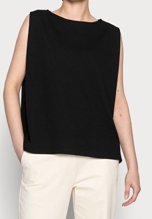 Person iført løs sort ærmeløs tekstureret top og off-white bukser med hånd i lommen mod ensfarvet baggrund.