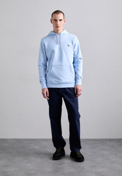 Felpa blu chiaro con tasca a marsupio e coulisse. Abbinata a pantaloni blu scuro e sneakers nere con dettagli verdi.