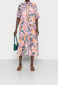 Vestido floral de color púrpura claro con mangas cortas, cuello tipo lazo y dobladillo con volantes, que presenta grandes flores y patrones de hojas. Sandalias naranjas y bolso verde.
