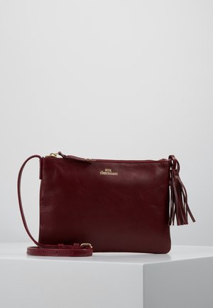 Ciemnoczerwona skórzana torebka na ramię z złotym zamkiem błyskawicznym, ozdobą z frędzli po boku oraz regulowanym paskiem, umieszczona na białej powierzchni na jednolitym szarym tle.