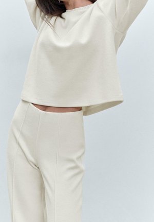 Femme portant un top court côtelé crème à manches longues et un pantalon large taille haute assorti, bras levés au-dessus de la tête.