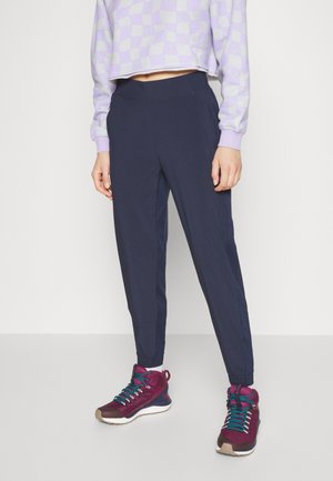 Kvinde i marineblå joggerbukser, lilla ternet cropped sweatshirt og lilla sportssko med turkise snørebånd stående på en ensfarvet hvid baggrund.