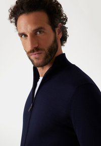 Maglione zip-up blu navy realizzato in un morbido tessuto testurizzato; presenta un colletto alto e dettagli di cucitura sottili. Indossato sopra una camicia bianca.