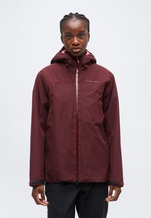Jeune personne portant une veste à capuche bordeaux avec une fermeture éclair sur le devant et un pantalon noir, debout devant un fond clair uni.