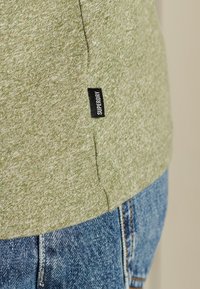 Grünes, strukturiertes Stoffhemd mit einem schwarzen "Superdry"-Etikett, kombiniert mit blauen Jeans. Das Hemd hat eine lockere Passform und seitliche Nahtdetails.
