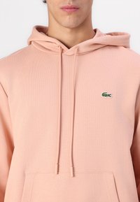 Sudadera de color rosa claro hecha de una tela suave, con un bolsillo canguro y un pequeño logo bordado en verde en la parte izquierda del pecho.
