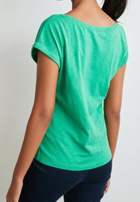 Camiseta de algodón verde con mangas enrolladas, escote redondeado y un corte holgado. La textura suave y la mezcla de tejidos crean un aspecto casual.