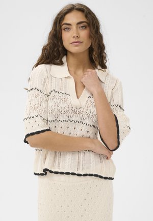 Naine pikkade lokkis juustega, kandes kreemjat kootud pluusi ja sobivat seelikut musta ääristusega, seistes ühe käega kaela juures.