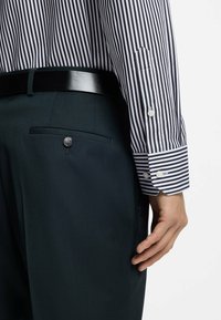 Gestreept overhemd met verticale strepen in marineblauw en wit; gecombineerd met donkergroene broek met een achterzak met knoop en een glanzende riem.