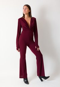 Tuta bordeaux con maniche lunghe, scollo a V profondo, design frontale annodato, silhouette aderente e gambe svasate. Indossata con stivali neri con tacco.