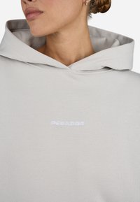 Hellgrauer Kapuzenpullover mit einer weichen Textur, Zugbandkapuze und einem weißen gestickten Logo auf der Brust. Einfaches und modernes Design.