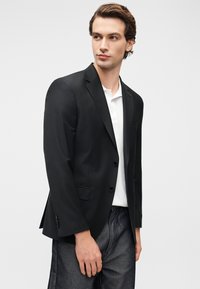 Μαύρο σακάκι με ανοιχτές πέτσες, δύο κουμπιά και τσέπη στο στήθος. Φορεμένο πάνω από λευκό πόλο, συνδυασμένο με σκουρόχρωμο τζιν παντελόνι.