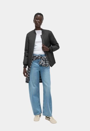 Veste bomber noire, t-shirt blanc, chemise à carreaux nouée à la taille, jeans larges bleu clair et baskets beiges.