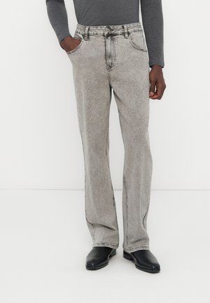 Άντρας που φοράει ανοιχτό γκρι τζιν παντελόνι με ίσια γραμμή, μακρυμάνικο μπλουζάκι σκούρου γκρι χρώματος και μαύρα δερμάτινα παπούτσια, όρθιος με το ένα χέρι στην τσέπη.