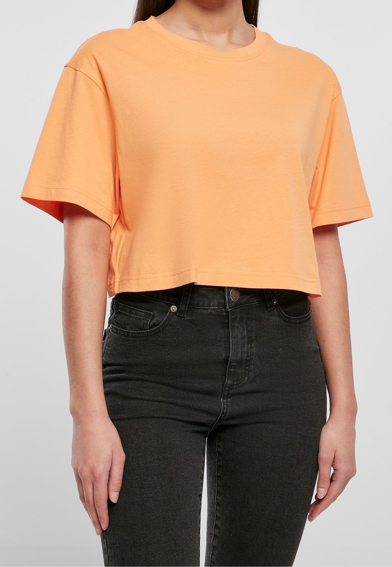 Femme portant un crop top orange ample à manches courtes associé à un jean noir taille haute sur un fond gris uni.