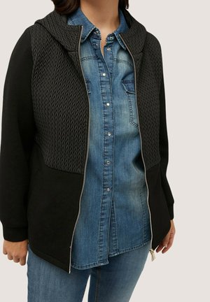 Kvinde iført en sort lynlåstrøje med teksturerede øverste paneler over en blå denimskjorte med knapper og blå jeans, stående mod en ensartet baggrund.