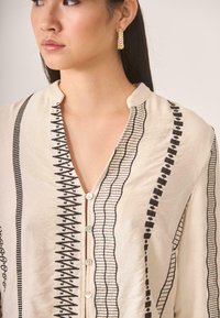 Blusa de color beige claro con intrincados bordados en negro, cuello alto y cierre de botones en la parte delantera. Textura suave con patrones detallados.