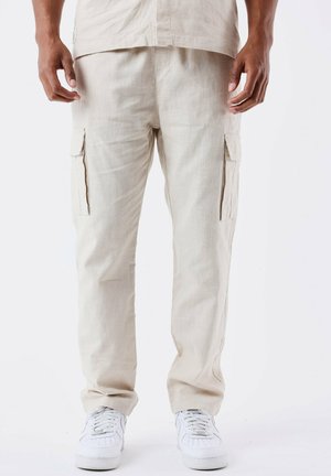 Pantalones cargo beige hechos de tela texturizada, con bolsillos laterales y un corte relajado, combinados con zapatillas blancas.
