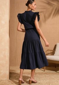Frocks albastru marin, cu straturi, mâneci volan, talie ajustată și fustă fluidă, completate de sandale cu toc înalt de culoare maro.