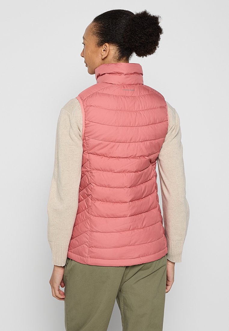 Gilet isolante rosa chiaro con colletto alto, design trapuntato e texture morbida; indossato sopra una maglietta a maniche lunghe color crema e pantaloni verdi.