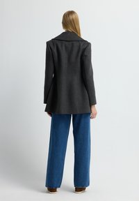 Blazer entallado gris oscuro con solapas anchas, hombros estructurados y espalda acampanada, combinado con jeans de pierna ancha azules.